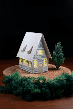 A small, detailed model house with illuminated windows stands on a round wooden platform. The house features a high peaked roof, decorative beams, and multiple windows. A tree is positioned beside the house, with dense green foliage surrounding the platform.