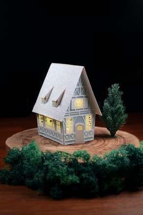 A small, detailed model house with illuminated windows stands on a round wooden platform. The house features a high peaked roof, decorative beams, and multiple windows. A tree is positioned beside the house, with dense green foliage surrounding the platform.