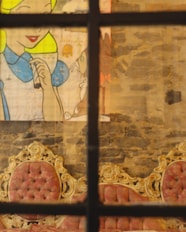 The image features a section of a wall with a painted cartoon-like character. This image includes a depiction of a figure with yellow hair and a blue outfit. In front of the painting are ornate chairs with red, tufted upholstery and elaborate gold detailing. The wall is textured with a rustic, exposed stone or brick look.