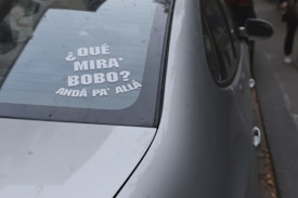 A car's rear window displays a message in Spanish, reading '¿Qué mirá' bobo? Andá pa' allá,' indicating a playful or confrontational tone. The silver color of the car is noticeable, and it is parked on the street with a few leaves scattered around.