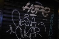 Graffiti on a dark metal shutter featuring the word 'HOPE' in large, bold letters at the top. Additional text includes the words 'O ESTA OKUR' and various other smaller writings in white and teal, scattered across the surface.