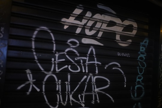 Graffiti on a dark metal shutter featuring the word 'HOPE' in large, bold letters at the top. Additional text includes the words 'O ESTA OKUR' and various other smaller writings in white and teal, scattered across the surface.