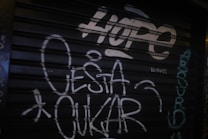 Graffiti on a dark metal shutter featuring the word 'HOPE' in large, bold letters at the top. Additional text includes the words 'O ESTA OKUR' and various other smaller writings in white and teal, scattered across the surface.
