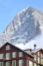 A towering, snow-capped mountain looms in the background with a clear blue sky above. In the foreground, a traditional wooden chalet with green window shutters and a red-brown facade is visible, with wisps of clouds behind it.