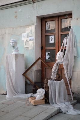 Various art pieces and sculptures are arranged artistically against the wall beside a wooden door. Plaster busts and canvas sketches are displayed, accompanied by draped white fabric and empty frames. The setting appears rustic with its weathered textures and subtle artistic flair.