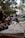 A detailed architectural model of a building is placed on a wooden surface in the foreground. The background features a group of people sitting and standing in a park-like setting, with trees and distant structures creating a blurred atmosphere.