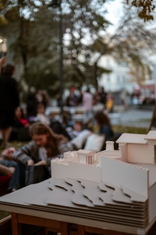 A detailed architectural model of a building is placed on a wooden surface in the foreground. The background features a group of people sitting and standing in a park-like setting, with trees and distant structures creating a blurred atmosphere.