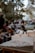 A detailed architectural model of a building is placed on a wooden surface in the foreground. The background features a group of people sitting and standing in a park-like setting, with trees and distant structures creating a blurred atmosphere.