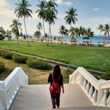 A person with a red backpack is walking down wide stone steps towards a grassy area lined with palm trees. Beyond the trees, there are several thatched-roof structures by a beach, with the ocean visible in the background. The scene is lit by soft daylight, indicating a clear and pleasant weather.