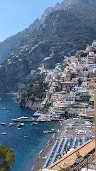 A picturesque coastal town with colorful buildings stacked on steep cliffs, overlooking a pristine blue sea. The beach is lined with rows of umbrellas and sunbeds, with boats scattered across the calm water. The mountainous backdrop adds to the dramatic scenery.