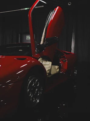 A sleek, red sports car with its vertical door open, showcasing the beige interior. The car is situated in a dark environment with a somewhat reflective black floor, and there is minimal lighting that highlights the contours and details of the vehicle.