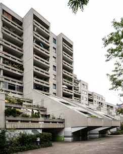 A modern multi-story apartment building with a unique architectural design features spacious balconies. The structure includes greenery such as plants and small trees on the terraces. The building is made of concrete with a series of angled ramps or overhangs that contribute to its distinct appearance. There is a paved area in front and trees on the sides add a touch of nature.