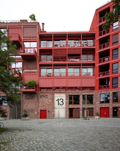 A modern apartment building with a red facade. The building features multiple balconies with railings and has a large number '13' displayed on the main entrance. The lower section of the building is constructed from brick, contrasting with the red upper stories. Trees and plants are visible around the building, adding a touch of greenery.