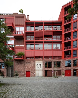A modern apartment building with a red facade. The building features multiple balconies with railings and has a large number '13' displayed on the main entrance. The lower section of the building is constructed from brick, contrasting with the red upper stories. Trees and plants are visible around the building, adding a touch of greenery.