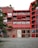 A modern apartment building with a red facade. The building features multiple balconies with railings and has a large number '13' displayed on the main entrance. The lower section of the building is constructed from brick, contrasting with the red upper stories. Trees and plants are visible around the building, adding a touch of greenery.