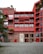 A modern apartment building with a red facade. The building features multiple balconies with railings and has a large number '13' displayed on the main entrance. The lower section of the building is constructed from brick, contrasting with the red upper stories. Trees and plants are visible around the building, adding a touch of greenery.