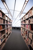 A modern library setting with two rows of bookshelves full of books leading to a reading area with several chairs. The ceiling features large skylights that allow natural light to illuminate the space.