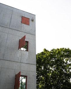 A modern concrete building facade features red-framed windows, some of which are open, allowing a view of lush green leaves from trees adjacent to the structure. The contrast between the hard, industrial feel of the concrete and the vibrant greenery creates a striking visual harmony.