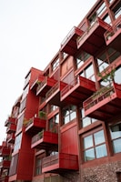 A multi-story building with a modern design featuring red protruding balconies, each adorned with potted plants. The structure includes large windows and a combination of red and rustic brown materials, creating a striking architectural aesthetic.