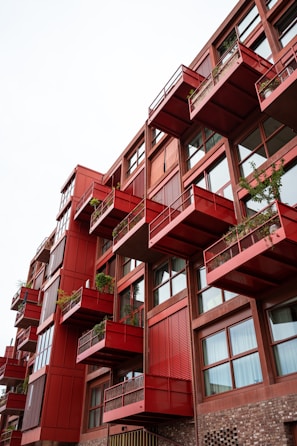 A multi-story building with a modern design featuring red protruding balconies, each adorned with potted plants. The structure includes large windows and a combination of red and rustic brown materials, creating a striking architectural aesthetic.