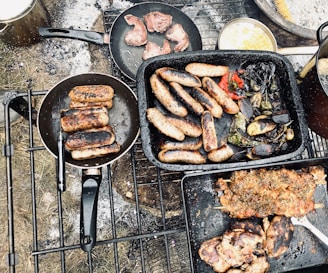A collection of cooked meats and vegetables on a grill. Two frying pans contain sausages, while another pan holds small pieces of meat. A roasting pan is filled with sausages, red peppers, and other grilled vegetables. Grilled meat skewers are placed in a tray beside some grilled eggplant.
