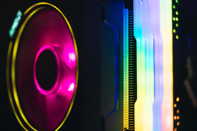 A close-up view of computer components featuring vibrant RGB lighting, including a glowing fan on the left and vertically aligned, luminous slots on the right. The lights create a spectrum of colors, blending from pink and purple to blue and yellow.