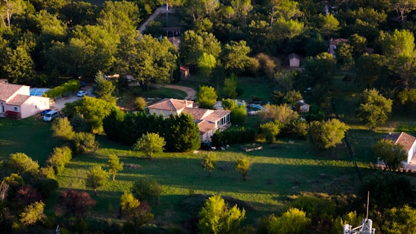 Aerial view of a rural area featuring several houses surrounded by lush greenery and trees. The landscape includes patches of grass, garden areas, and a few parked cars along narrow dirt roads. The sun casts soft shadows, indicating early morning or late afternoon.