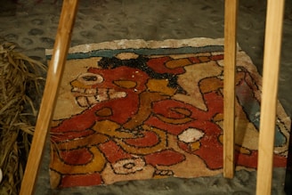 A rustic, colorful sand painting or mural is laid out on a stone floor, featuring intricate designs and abstract shapes in red, yellow, and black with a hint of blue. Two wooden poles are positioned above it, and dry straw is visible on one side. The artwork seems to depict a stylized animal or mythical creature.