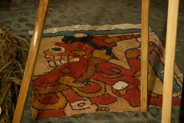 A rustic, colorful sand painting or mural is laid out on a stone floor, featuring intricate designs and abstract shapes in red, yellow, and black with a hint of blue. Two wooden poles are positioned above it, and dry straw is visible on one side. The artwork seems to depict a stylized animal or mythical creature.