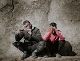 Two men are seated with their backs against a rough, textured wall. The man on the left is wearing a dark jacket and pants, appearing disheveled and thoughtful or concerned as he looks at an object he is holding. The man on the right is wearing a red sports jacket with white stripes and appears to be covering his face or eyes with his hands, possibly in distress or despair.