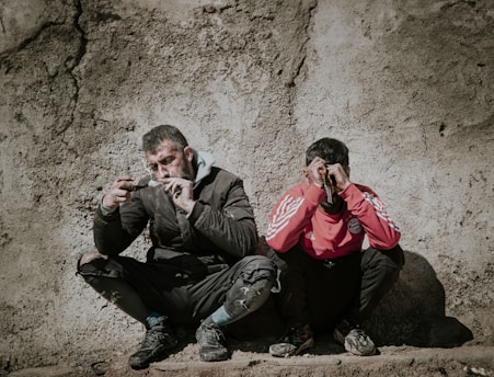 Two men are seated with their backs against a rough, textured wall. The man on the left is wearing a dark jacket and pants, appearing disheveled and thoughtful or concerned as he looks at an object he is holding. The man on the right is wearing a red sports jacket with white stripes and appears to be covering his face or eyes with his hands, possibly in distress or despair.