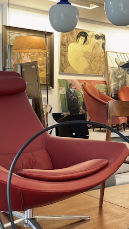 A vintage-inspired room features an array of stylish furniture and artwork. A prominent red leather lounge chair with curved lines is positioned in the foreground. The background showcases multiple large framed paintings, including a traditional Asian portrait. Various other pieces of furniture and decorative lamps add to the sophisticated ambiance.