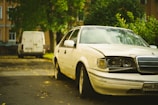 A white sedan with noticeable front-end damage is parked on a street next to some green foliage. In the background, a white van is parked further down the road. The setting appears to be residential, with trees and buildings lining the street.