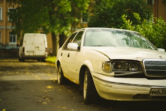 A white sedan with noticeable front-end damage is parked on a street next to some green foliage. In the background, a white van is parked further down the road. The setting appears to be residential, with trees and buildings lining the street.