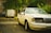 A white sedan with noticeable front-end damage is parked on a street next to some green foliage. In the background, a white van is parked further down the road. The setting appears to be residential, with trees and buildings lining the street.