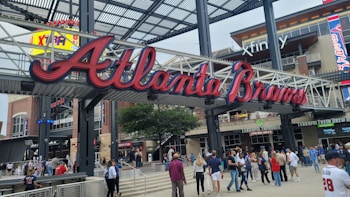A large sign with the words 'Atlanta Braves' in red, cursive letters is suspended from a metal framework. Below, a bustling crowd of people moves about, some wearing sports apparel. The background features a multistory building with signage, including 'Xfinity' and 'Terrapin Taproom.'