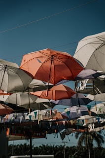 A collection of colorful umbrellas hangs suspended in the air, creating an artistic canopy above. The umbrellas vary in colors, with shades of orange, white, and purple prominently visible. Beneath the umbrellas, colorful triangular flags add to the festive vibe. Palm trees are visible in the background under the clear blue sky.