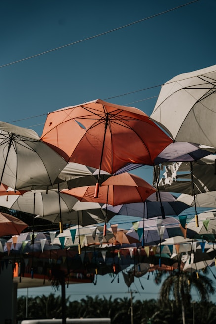 A collection of colorful umbrellas hangs suspended in the air, creating an artistic canopy above. The umbrellas vary in colors, with shades of orange, white, and purple prominently visible. Beneath the umbrellas, colorful triangular flags add to the festive vibe. Palm trees are visible in the background under the clear blue sky.
