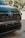 Close-up of the front grill of a black Toyota vehicle, showcasing the emblem prominently in a well-lit outdoor setting. The background is blurred with greenery and hints of signage visible.