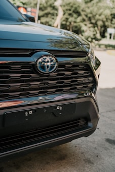 Close-up of the front grill of a black Toyota vehicle, showcasing the emblem prominently in a well-lit outdoor setting. The background is blurred with greenery and hints of signage visible.