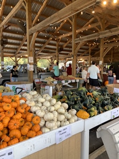 A rustic farmer’s market with wooden beams and string lights features a variety of produce. In the foreground, small orange, white, and green pumpkins are displayed. People are browsing the stalls under a roofed area with a vintage truck visible on the left.
