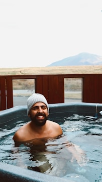 A man wearing a beanie is happily soaking in a hot tub outdoors, with a mountainous landscape in the background. The wooden railing around the tub frames the scene, and the man is smiling broadly, exuding warmth and relaxation.