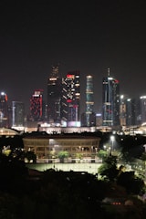 A nighttime cityscape featuring modern skyscrapers illuminated with colorful lights in the background. In the foreground, a large, well-lit building with surrounding green spaces and sports courts is visible.