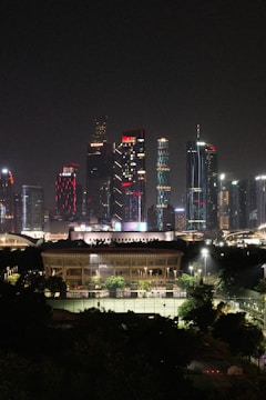 A nighttime cityscape featuring modern skyscrapers illuminated with colorful lights in the background. In the foreground, a large, well-lit building with surrounding green spaces and sports courts is visible.