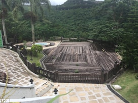 A large wooden deck surrounded by lush greenery overlooks a verdant hillside. Tall palm trees stand by the side, and a circular stone pathway leads to a small round pool. The scene is peaceful and secluded, suggesting a spot for relaxation amidst nature.