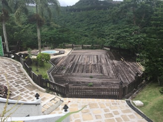 A large wooden deck surrounded by lush greenery overlooks a verdant hillside. Tall palm trees stand by the side, and a circular stone pathway leads to a small round pool. The scene is peaceful and secluded, suggesting a spot for relaxation amidst nature.