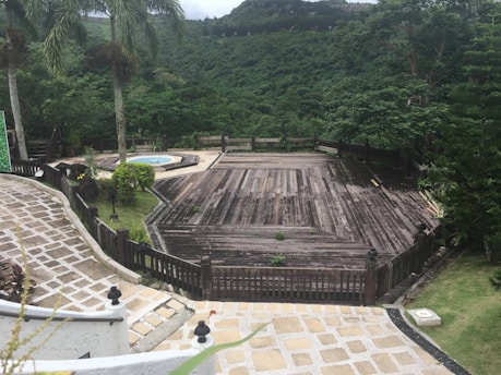 A large wooden deck surrounded by lush greenery overlooks a verdant hillside. Tall palm trees stand by the side, and a circular stone pathway leads to a small round pool. The scene is peaceful and secluded, suggesting a spot for relaxation amidst nature.