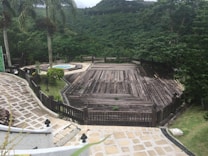 A large wooden deck surrounded by lush greenery overlooks a verdant hillside. Tall palm trees stand by the side, and a circular stone pathway leads to a small round pool. The scene is peaceful and secluded, suggesting a spot for relaxation amidst nature.