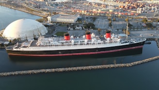 An aerial view of a large, historic ocean liner with three distinct red and black smokestacks, docked in a port. A geodesic dome structure is visible on the land nearby, along with parking lots, trees, and other port infrastructure. The calm water reflects the colors of the ship and the surrounding environment.