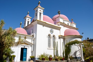 A church with pink domes and white walls is set against a clear blue sky. The structure features several crosses atop its domes and multiple arched windows with decorative designs. In the foreground, there are several tall cacti and other desert plants.